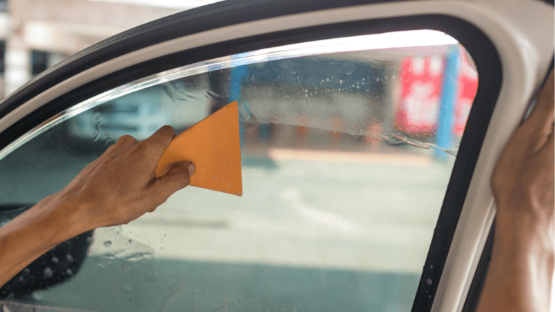 A man is smoothing the car window film with a plastic scraper.