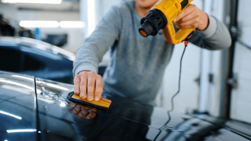 A man is installing a vinyl wrap on the car with a plastic scraper and a heat gun.