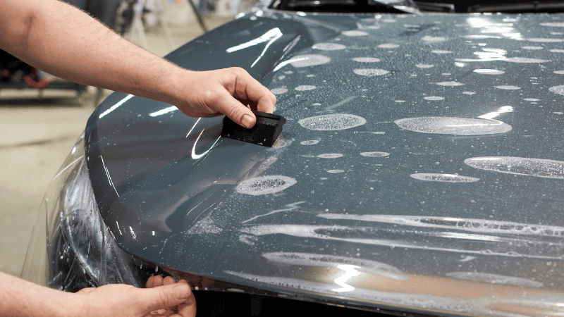 A man is installing a vinyl wrap on the car with a plastic scraper.
